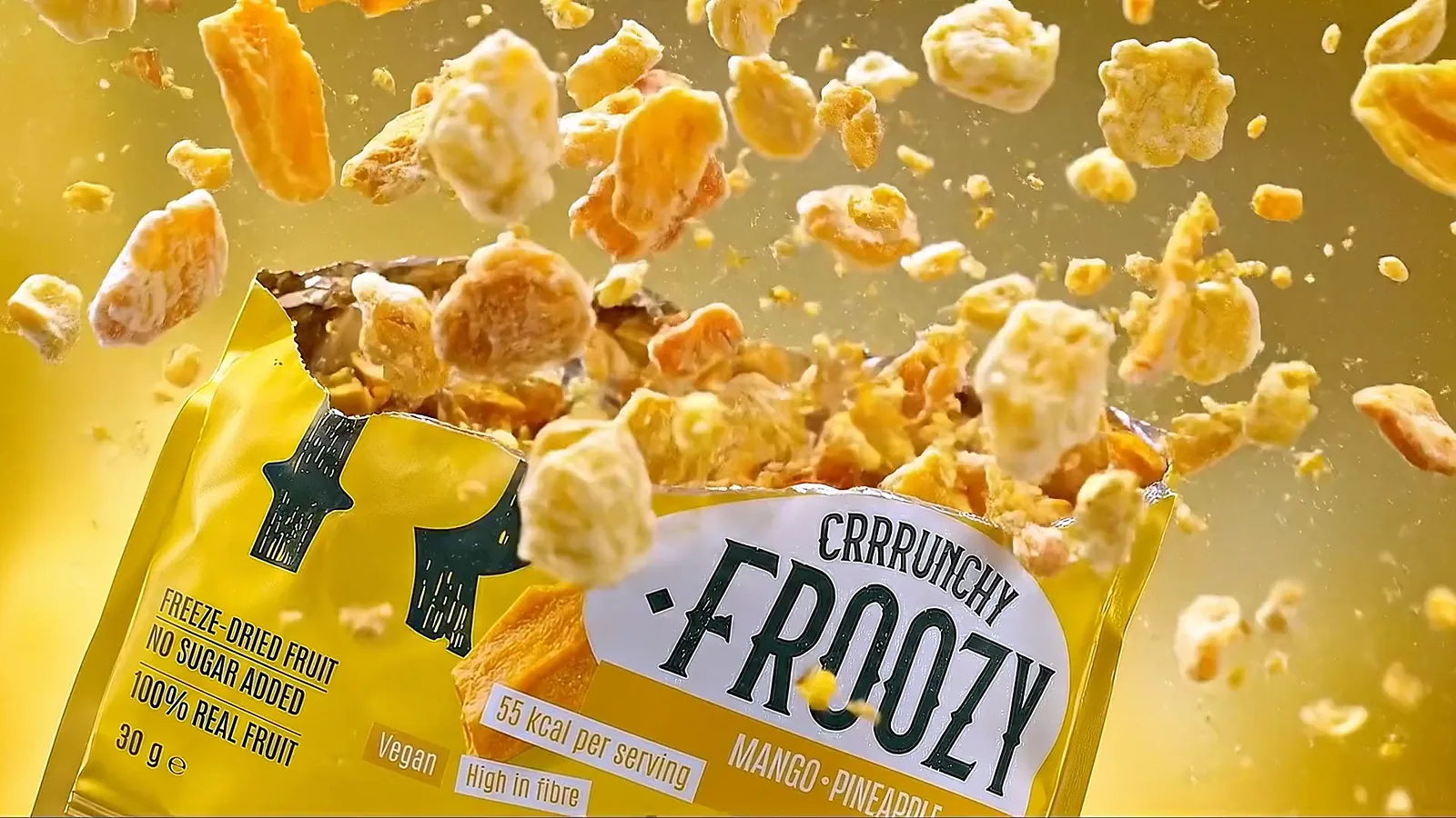 Een geopend zakje Froozy Mango · Pineapple waar gevriesdroogd fruit uit omhoog spat, tegen een goudgele achtergrond